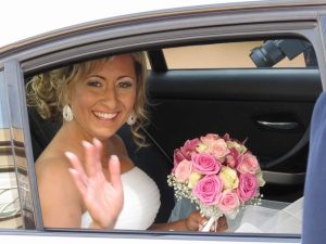 Novia feliz saludando desde el coche con ramo de flores en su boda