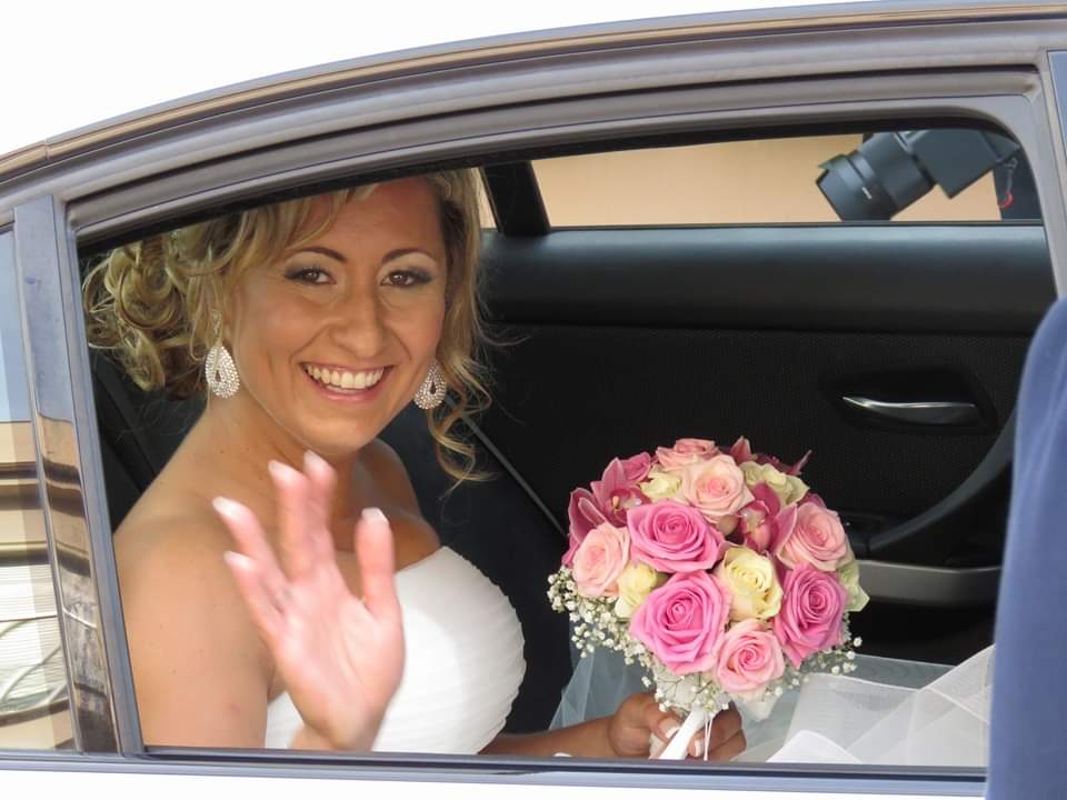 Novia feliz saludando desde el coche con ramo de flores en su boda